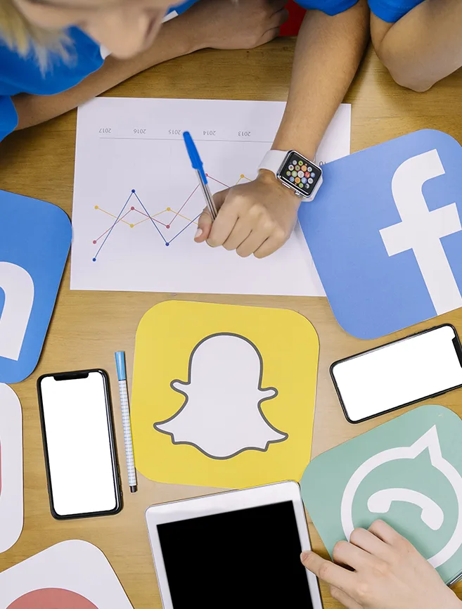 A top-down view of a table with various social media platform icons, including Snapchat, Facebook, WhatsApp, and LinkedIn, along with smartphones and a tablet.